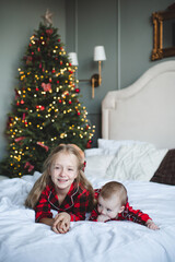 Happy children lying in bed and having fun over Christmas tree and glowing lights wearing red pajamas in room at home. Winter holiday season. Childhood.