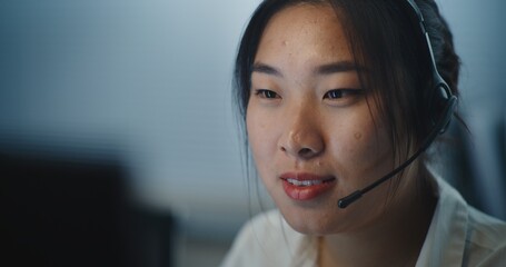 Call center office: Close up portrait of Asian female helpdesk specialist talking with client on headset, looking at PC screen, providing online customer service. Technical support operator working.