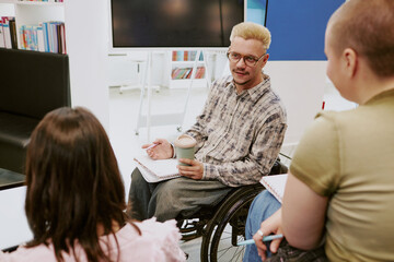 Wheelchair user holding beverage and engaging in group discussion with colleagues in a modern office setting using notebooks and tablets