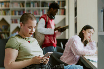 Students studying in a library setting with bookshelves in background, using smartphones and reading material. Scene captures focus and concentration while engaging with digital and printed resources