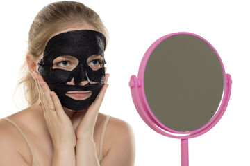 Beautiful young woman puts a facial black paper mask on her face. Skin care and treatment, spa, natural beauty and cosmetology concept