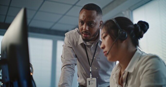 Call center office: African American technical support specialist helps Asian colleague in headset. Two multiethnic hotline operators working in online customer assistance service, using computer.