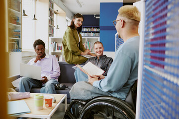 Diverse team members collaborating in modern office space, sitting comfortably, and using laptops and books. Inclusivity highlighted with wheelchair accessibility