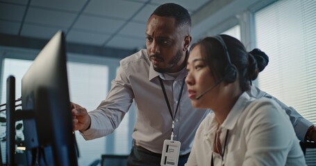 Call center office: African American technical support specialist helps Asian colleague in headset....
