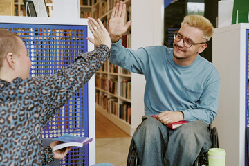 Two friends sharing high five in library surrounded by shelves with books while holding their own...