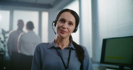 Call center office: Close up portrait of friendly Caucasian female technical customer support specialist wearing headset smiling, looking at camera. Team of hotline operators standing in background.