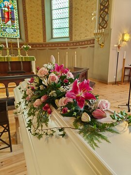 White coffin adorned with a beautiful floral arrangement of pink and red flowers, set in a solemn interior with candles in the background, creating a respectful funeral scene