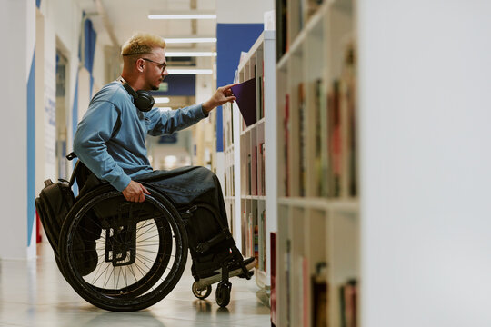 Young male student in wheelchair browsing library shelves for books while wearing headphones and casual attire. Focused individual engaging in academic environment with assistive device