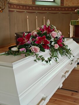 White coffin adorned with a beautiful floral arrangement of pink and red flowers, set in a solemn interior with candles in the background, creating a respectful funeral scene