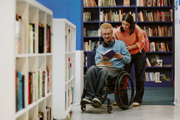 Young man in a wheelchair reading book with assistance of young woman in library filled with...