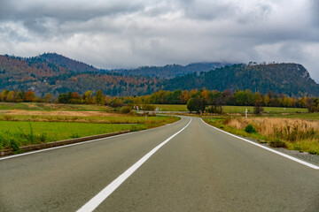 Road trip. Empty winding countryside road between fields in autumn time. Beautiful mountain slopes covered with colourful forest. Cloudy weather 
