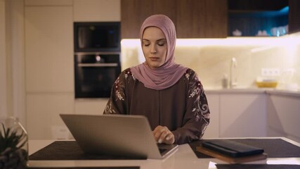 Arab female student in headscarf typing on laptop at home, studying online