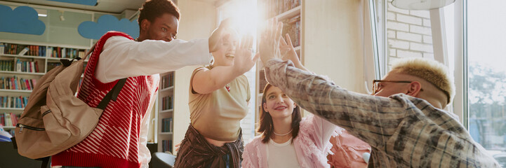 Group of diverse students high-fiving and smiling inside library filled with bookshelves,...