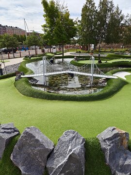 Miniature golf course with a small bridge over a pond, surrounded by artificial grass, rocks, and greenery. A fun outdoor activity setting