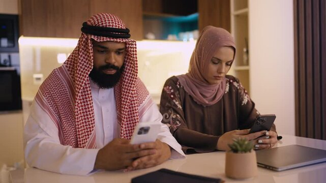 Arab couple in traditional clothing scrolling on their phones, not communicating
