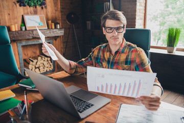 Photo of handsome young man wear shirt sit table hold read paper analyzing report stats workplace...
