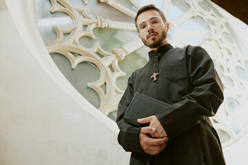 Low angle portrait of young Caucasian priest holding Bible looking at camera while standing next to...