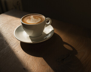 A coffee in a white cup on wooden table, resting in the warmth of the sun. There is detailed latte art in the steamed milk. The coffee casts a warm shadow.