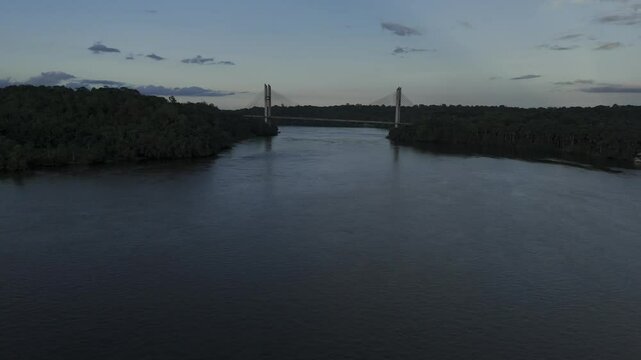 Drone flies at sunset along Oiapoque river toward south side of bridge between Brazil and French Guayana near Oiapoque, Amap&aacute;, Brazil