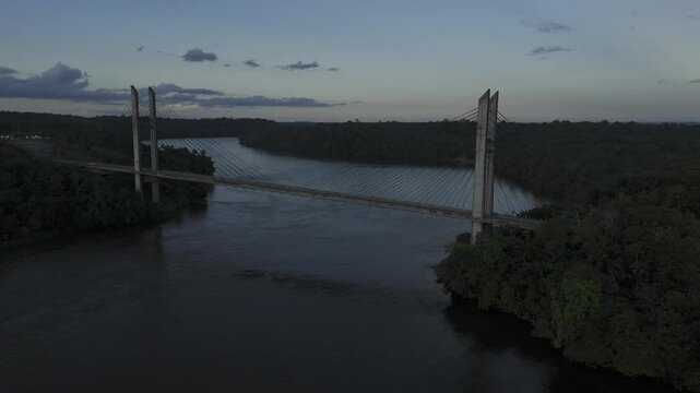 Drone hovers high over river overlooking suspension bridge during sunset