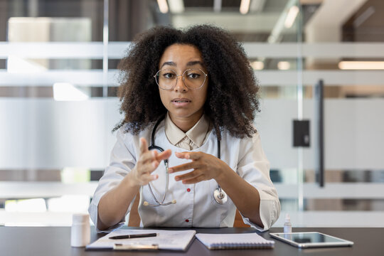 A young African American female doctor in a white coat and glasses is sitting at a desk in a hospital office, talking and consulting at the camera while gesturing with her hands