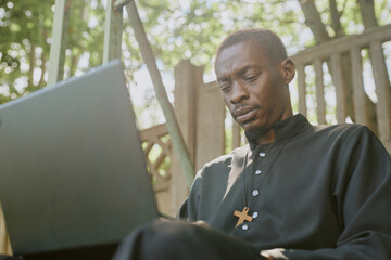 Low angle shot of African American priest dressed in black robe with wooden cross on chest sitting on bench and using laptop