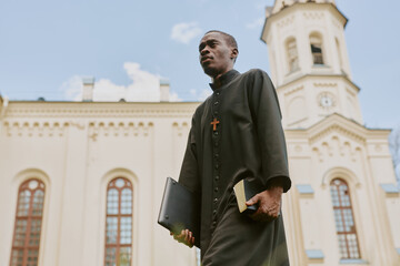 Low angle shot of African American priest in black robe with big wooden cross carrying laptop and...