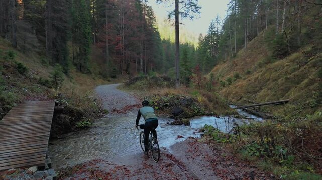 Cyclist on gravel bike crosses cold mountain river along forest trail in mountains, choosing challenge of water instead of wooden bridge nearby. Autumn colors surround. Bicyclist rides across creek. 