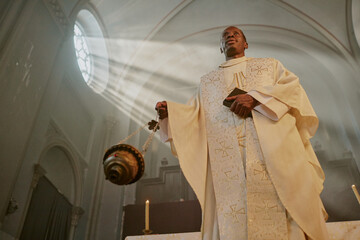 Low angle shot of mature African American priest in golden white robe swinging golden censer while...
