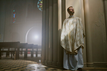 Young Caucasian priest in white robe leaning on majestic church wall before service
