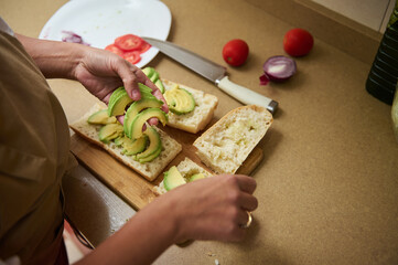 Person preparing a healthy avocado sandwich with fresh ingredients on a wooden cutting board in a cozy kitchen. The scene suggests a sense of homemade cooking and healthy eating lifestyle.