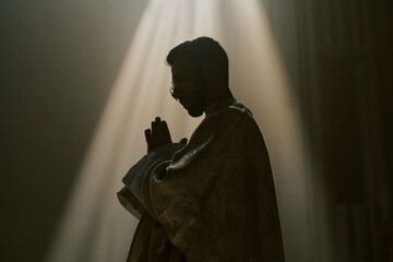 Low angle shot of unrecognizable priest clasped hands during praying in church illuminated with golden sunrays