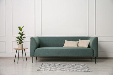 Stylish sofa, pillows and side table with houseplant near white wall indoors