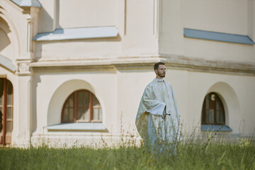 Caucasian priest in golden white robe walking slowly through yard with fresh cut grass