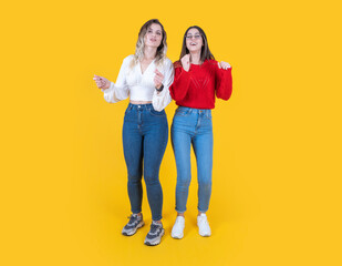 Fototapeta premium Energetic young two girls dancing, full body view caucasian 20s energetic young two girls dancing side by side waving hands in studio yellow background. Smiling happy two women listen music.