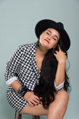 Studio portrait of a beautiful woman sitting on a wooden stool posing for a photo wearing a black hat and wearing black and white clothes.