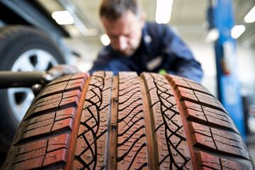 Mechanic Inspecting Car Tire in Auto Repair Shop. A close-up of a car tire with detailed tread patterns in focus, while a mechanic works in the background. 