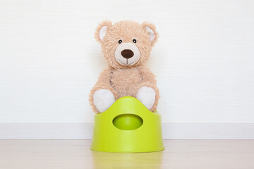 Smiling brown soft teddy bear sitting on baby potty on wooden home floor at light gray wall background. Closeup. Front view.