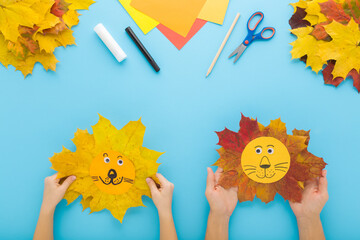 Mother and child hands creating lion head shapes from colorful maple leaves and paper. Blue table...