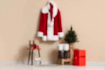Santa Claus suit on hanger with gift boxes, fir tree and boots near beige wall. Blurred view