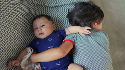 Baby lying next to older brother on a couch, both resting peacefully, baby smiling and stretching arms, enjoying a moment of comfort and connection