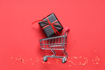 Mini shopping cart with gift box and confetti on red background. Black Friday sale. Top view