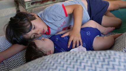 Older brother lying beside baby sibling on a couch, gently patting the baby’s belly, both...