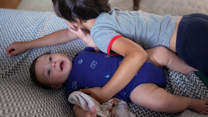 Older brother lying beside baby sibling on a couch, gently patting the baby’s belly, both...