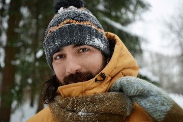 Close up portrait of bearded young man covered in snow looking at camera in winter forest with...