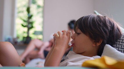 Young boy drinking water from a glass, relaxing on a couch in a calm home environment, with mother blurred in the background, quiet and reflective family moment