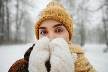 Close up portrait of young woman in winter outdoors looking at camera and blowing on hands with white fluffy mittens