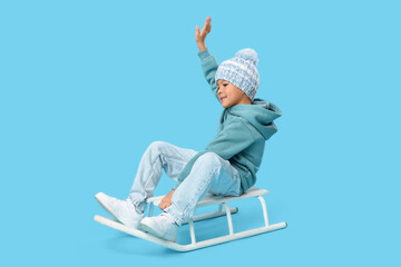 Cute little Asian boy in stylish knitted hat sitting on sledge against blue background