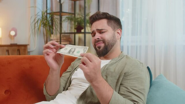 Poor Caucasian young man upset over insufficient amount of money, counting dollar banknotes. Financial crisis. Bankruptcy. Poverty and destitution. Upset adult guy sitting on sofa couch at home.