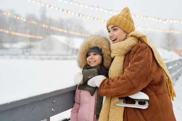 Candid shot of excited mother and daughter watching skating rink in winter park with twinkling garlands and snow falling copy space